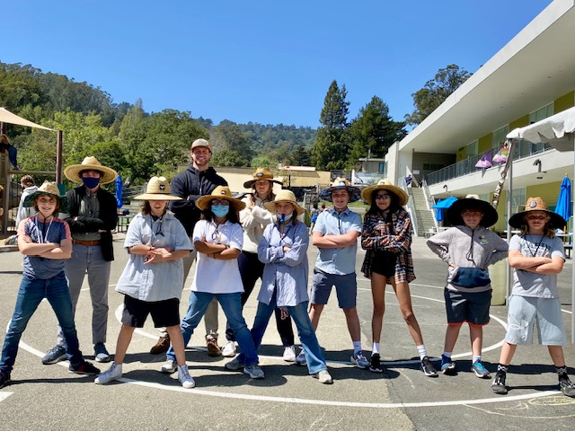 A group of students and adults wearing hats and posing together in a sunny school courtyard